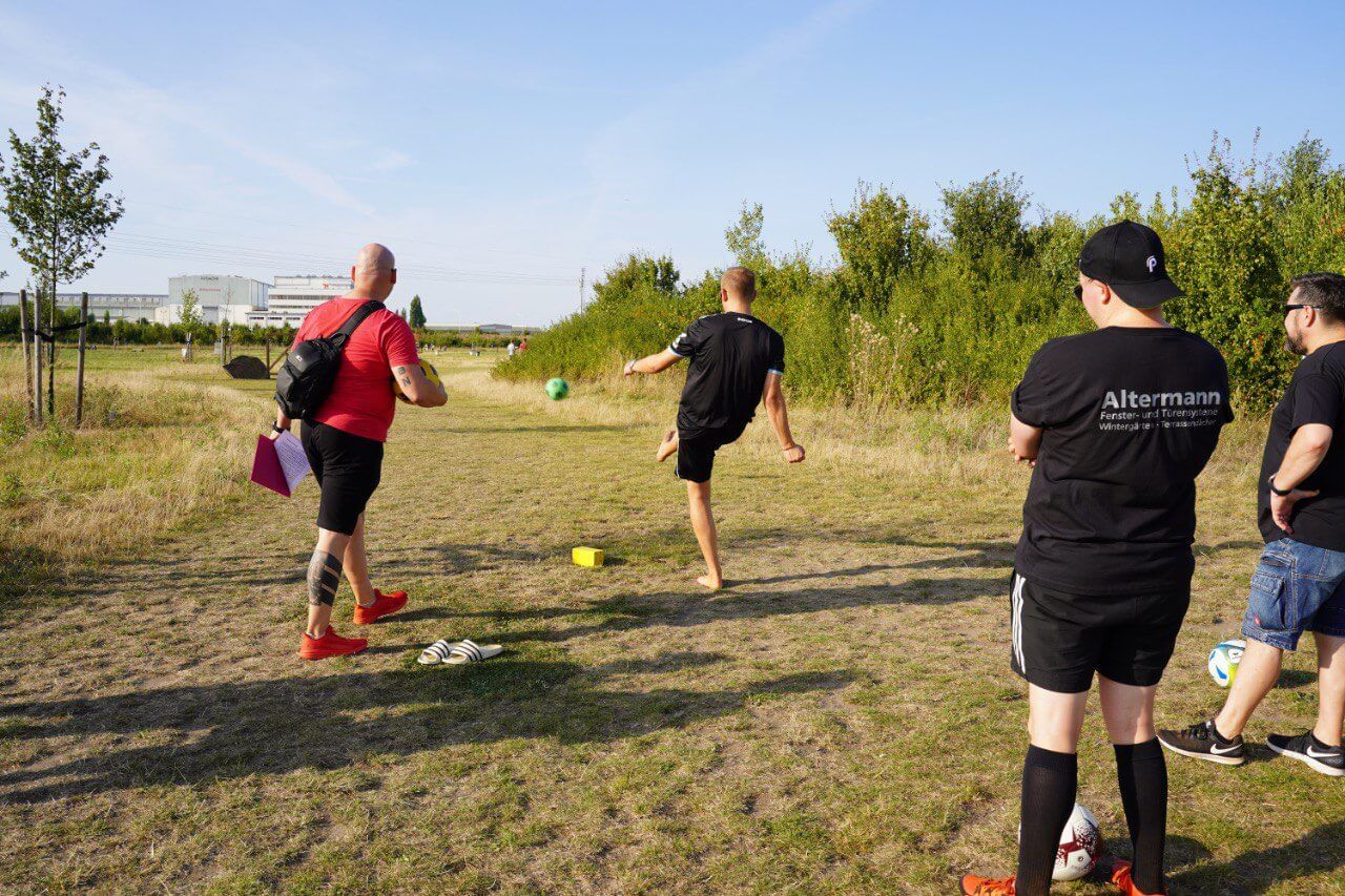 Eine Gruppe in Team-Shirts schaut zu, wie ein Spieler den Ball über eine Fußballgolf-Bahn schießt